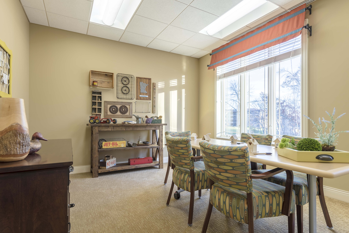 A light room with a big window furnished with a dining table and chairs, a chest of drawers, and shelving with framed pictures on the facing wall.